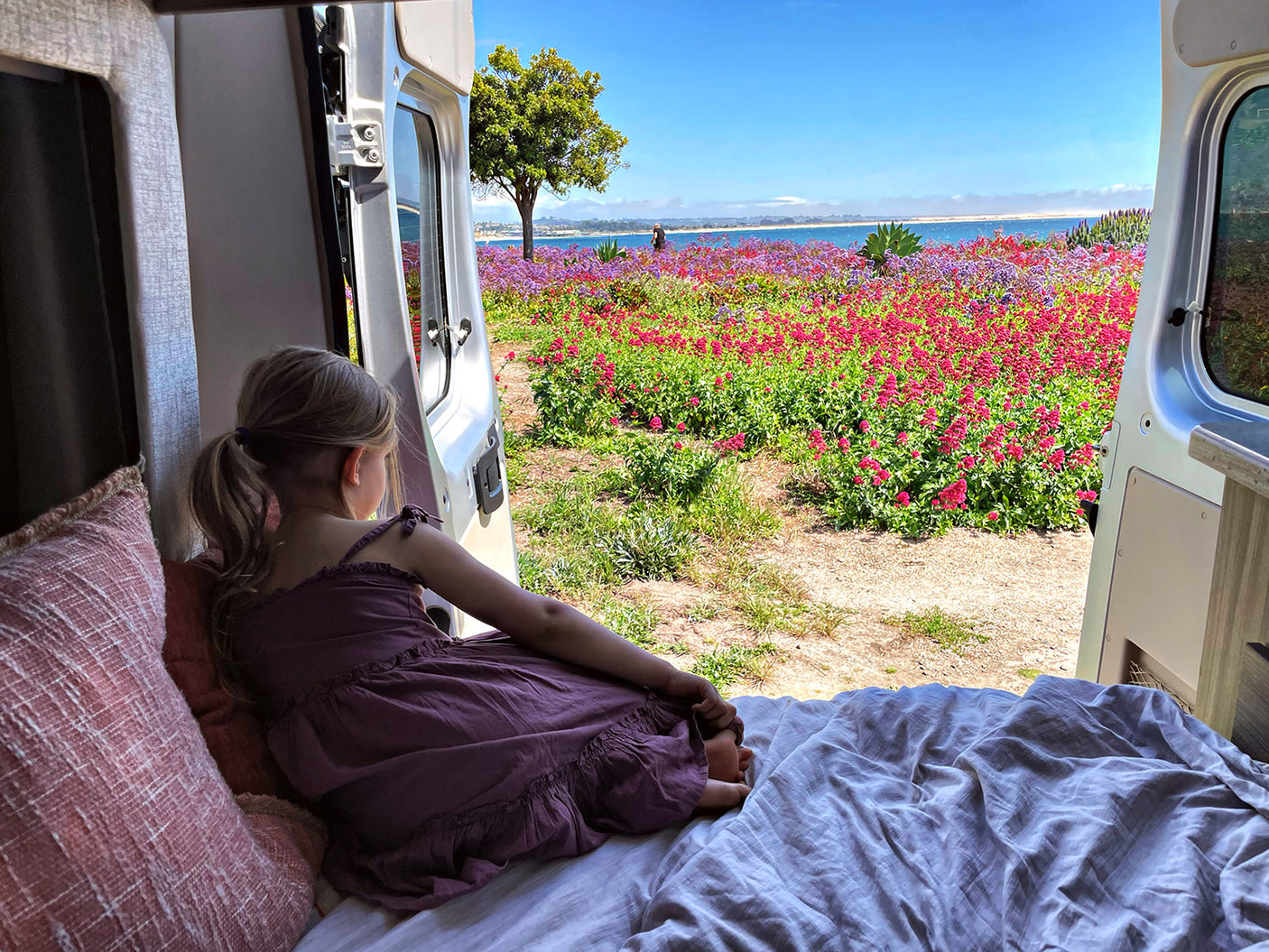 RV rental adventure: Young girl in a cozy van, enjoying vibrant flowers & ocean view from the open back doors.