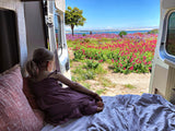 RV rental adventure: Young girl in a cozy van, enjoying vibrant flowers & ocean view from the open back doors.