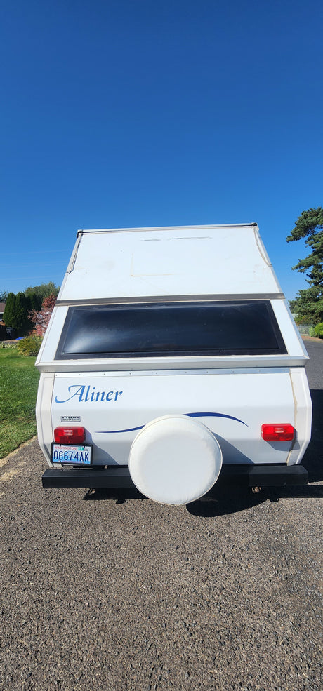 A-liner A-frame camper RV rental with WA plate 0G674AK & spare tire, parked on gravel under clear blue sky.
