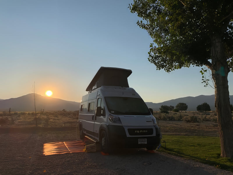 An RV rental camper van is parked at sunset with mountains behind. A large tree is on the right, and a mat catches light. — photo 15