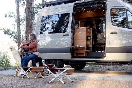 A joyful couple relaxes outside their modern RV rental van with its side door open, revealing a cozy interior.
