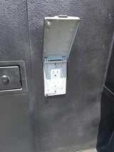 RV rental outdoor weatherproof outlet: A white 3-prong receptacle with an open gray cover on a dark, textured RV exterior.