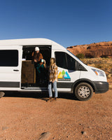 Two women exchange a mug by their white Andervans RV rental, ready for adventure. Ideal for outdoor travel & van life.