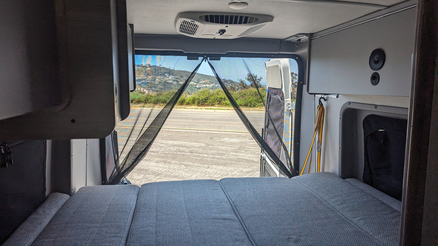 Inside an RV rental: a cozy bed faces open rear doors with a bug screen, revealing a road and hills. Ready for travel.