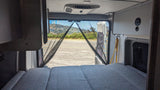 Inside an RV rental: a cozy bed faces open rear doors with a bug screen, revealing a road and hills. Ready for travel.