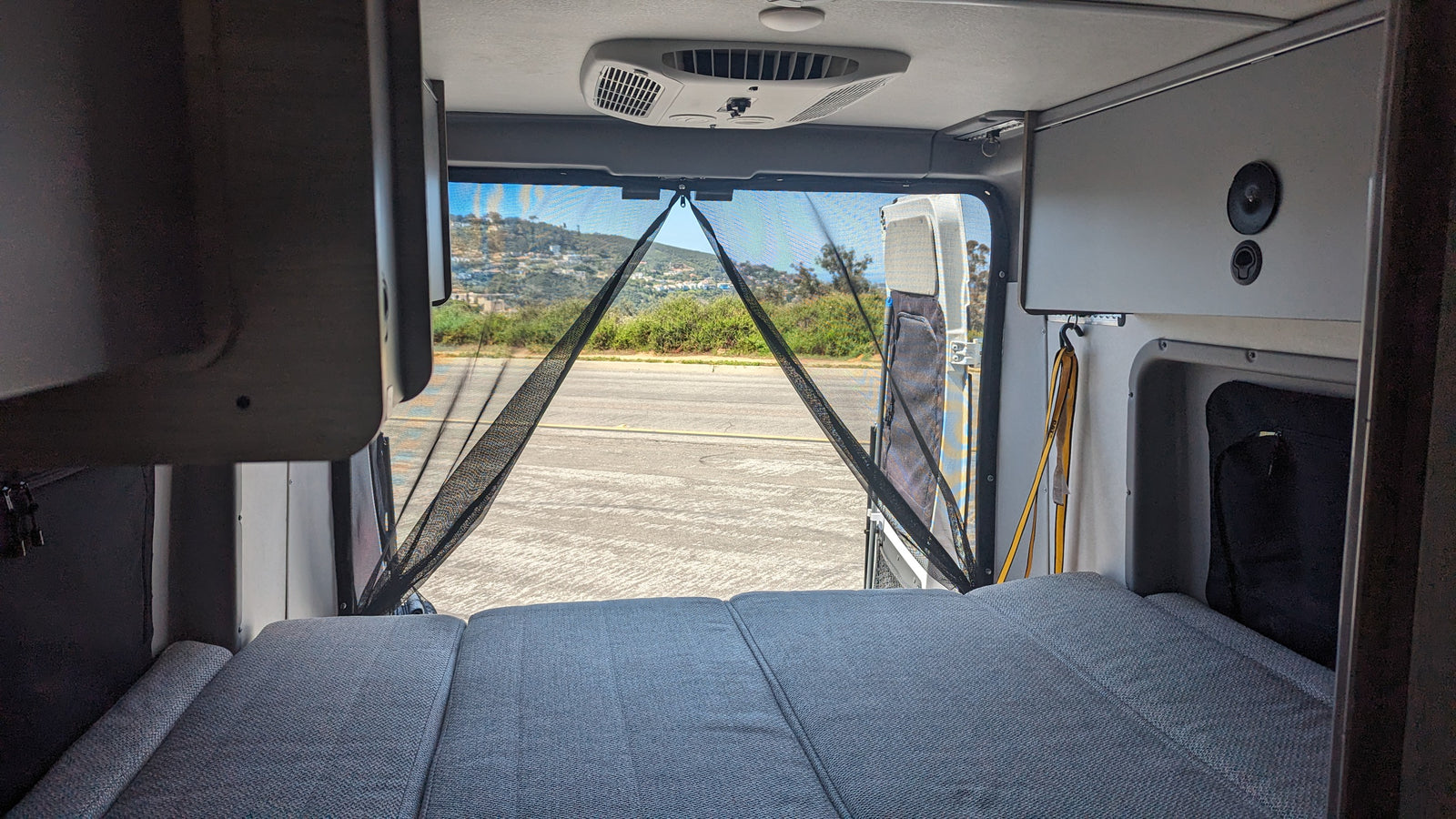 Inside an RV rental: a cozy bed faces open rear doors with a bug screen, revealing a road and hills. Ready for travel.