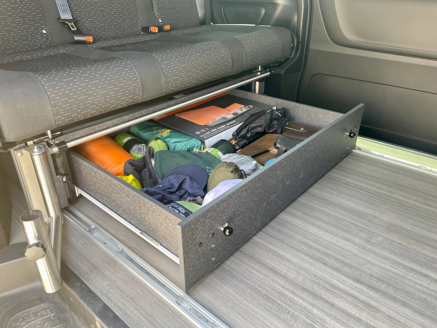 RV rental convenience: Organized pull-out storage drawer under a camper van seat, filled with camping gear.