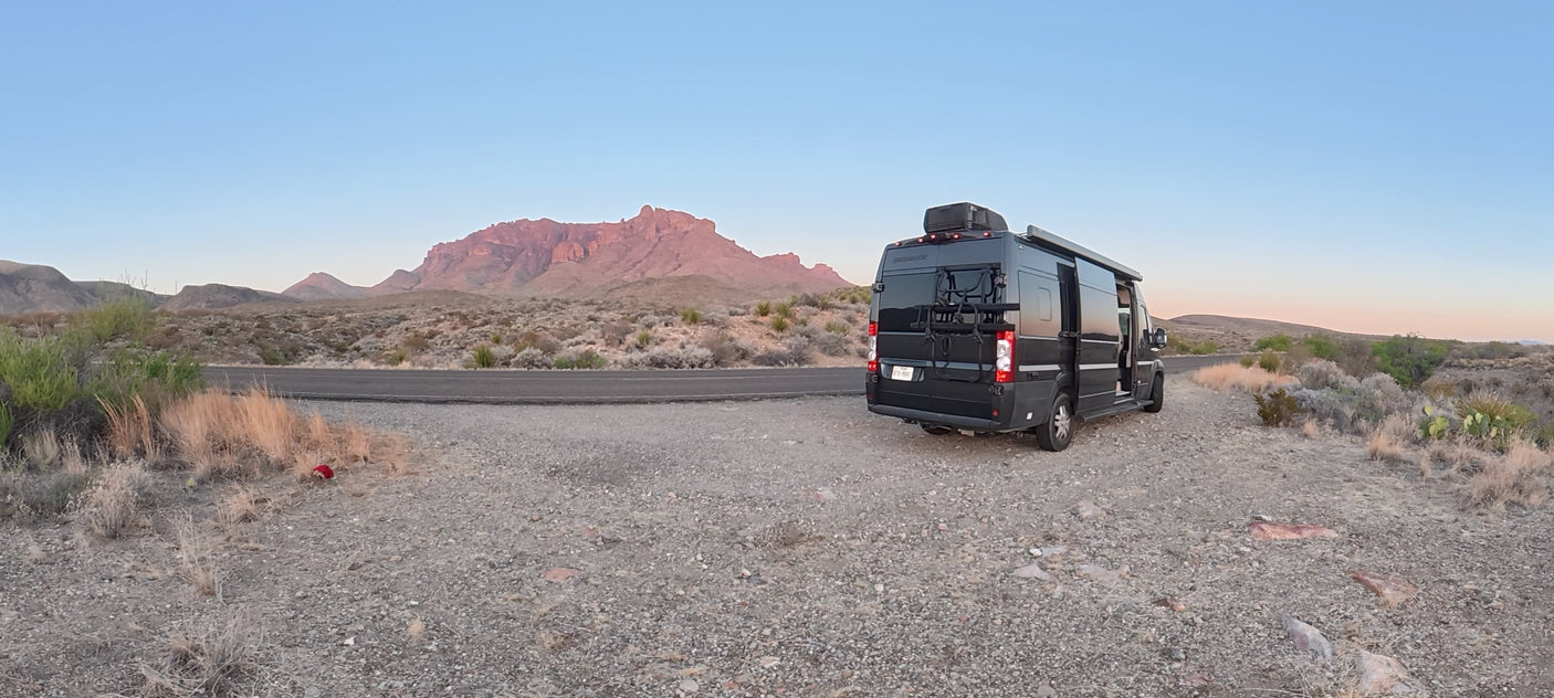 A dark RV rental is parked on a gravel shoulder overlooking a desert landscape with large, reddish mountains under a soft, clear sky.