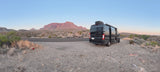 A dark RV rental is parked on a gravel shoulder overlooking a desert landscape with large, reddish mountains under a soft, clear sky.
