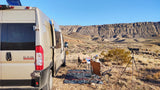 Enjoy a scenic RV rental adventure! A tan camper van is parked in a desert landscape with a camp setup & spotting scope.