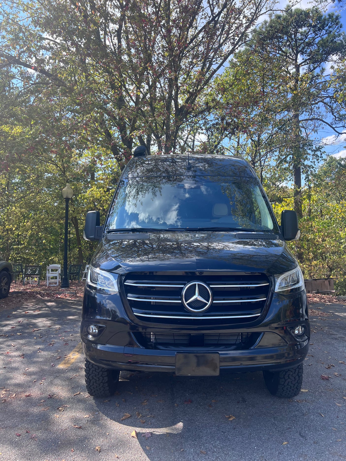 Black Mercedes-Benz Sprinter van, ideal for rv rental, parked on asphalt with a backdrop of autumn trees.