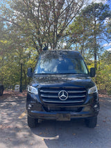 Black Mercedes-Benz Sprinter van, ideal for rv rental, parked on asphalt with a backdrop of autumn trees.