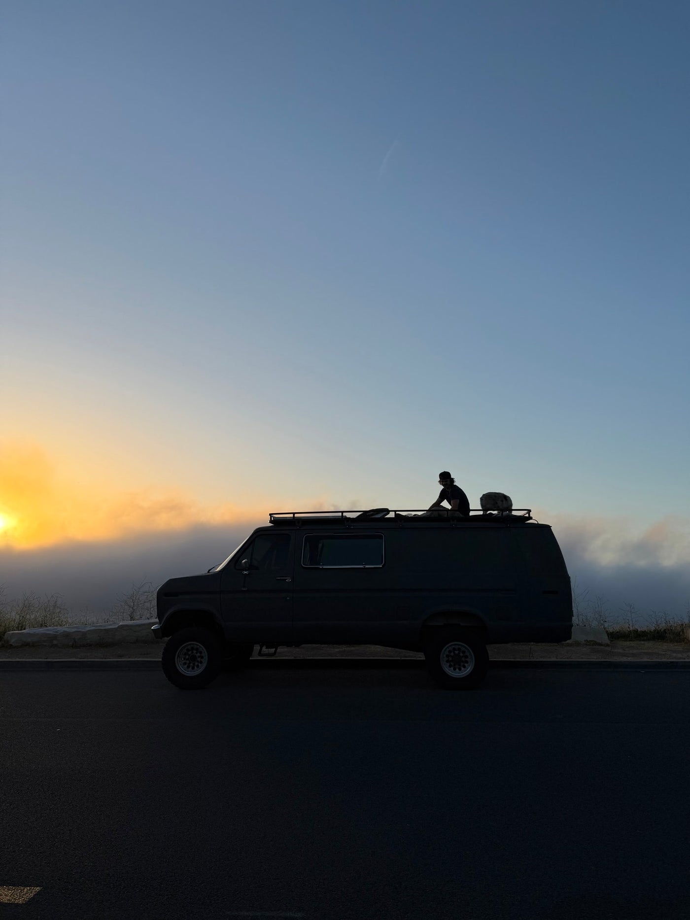 An RV rental, a dark camper van, is parked. A person sits silhouetted on its roof, watching a golden, cloudy sunrise. — photo 9