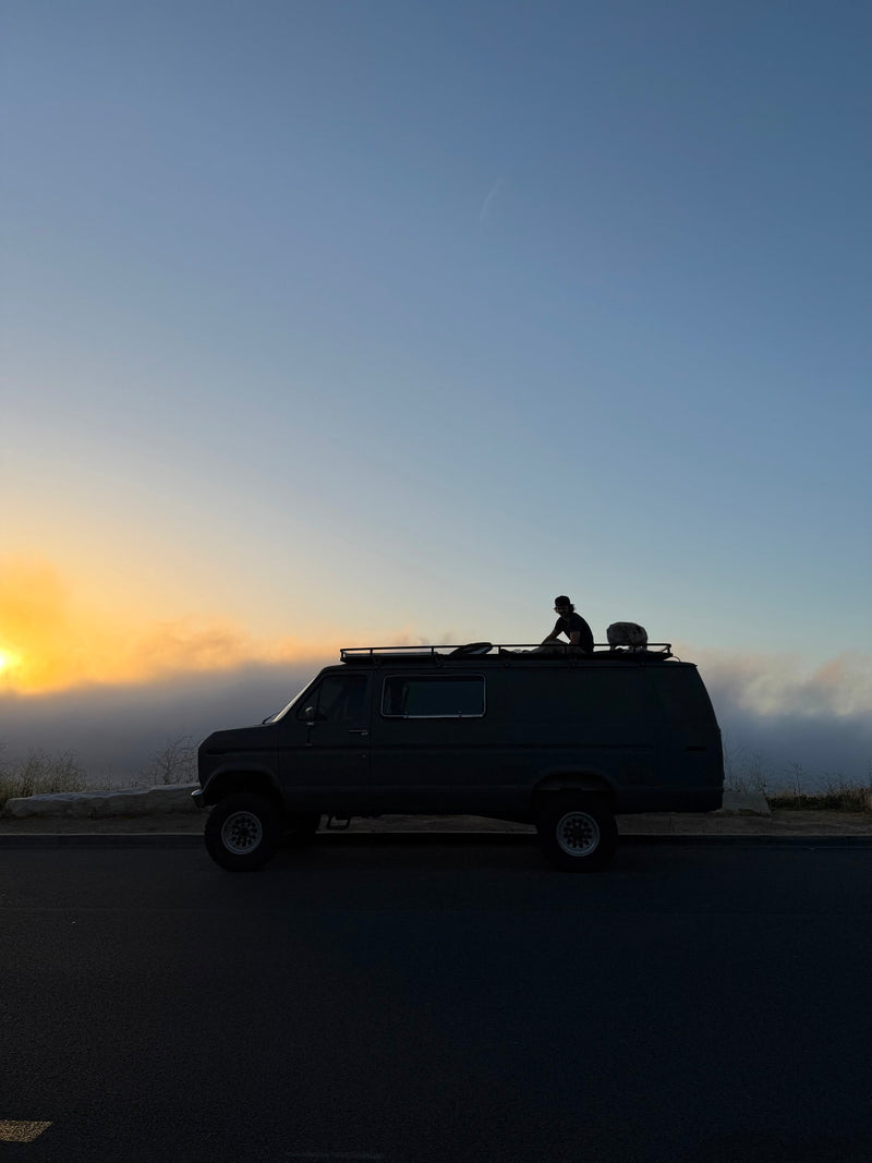 An RV rental, a dark camper van, is parked. A person sits silhouetted on its roof, watching a golden, cloudy sunrise. — photo 9