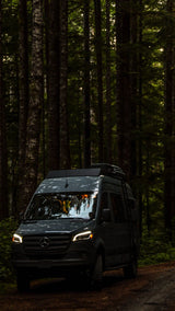 A dark grey Mercedes Sprinter van, headlights on, on a dirt road, amidst tall trees in a dim forest. Great RV rental.