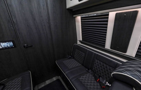 Modern RV rental interior with dark wood walls, quilted leather seats, a control panel, and window blinds.