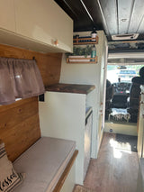 Bright rv rental interior featuring sage green cabinets, wood walls, and a cozy bench, with a view to the driver's cab.
