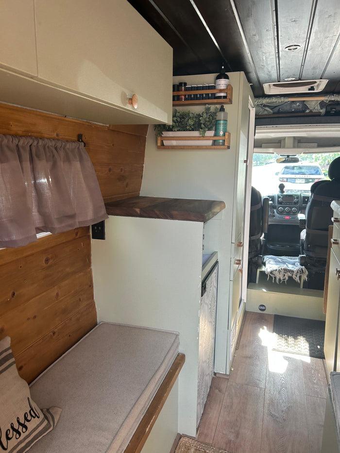 Bright rv rental interior featuring sage green cabinets, wood walls, and a cozy bench, with a view to the driver's cab.