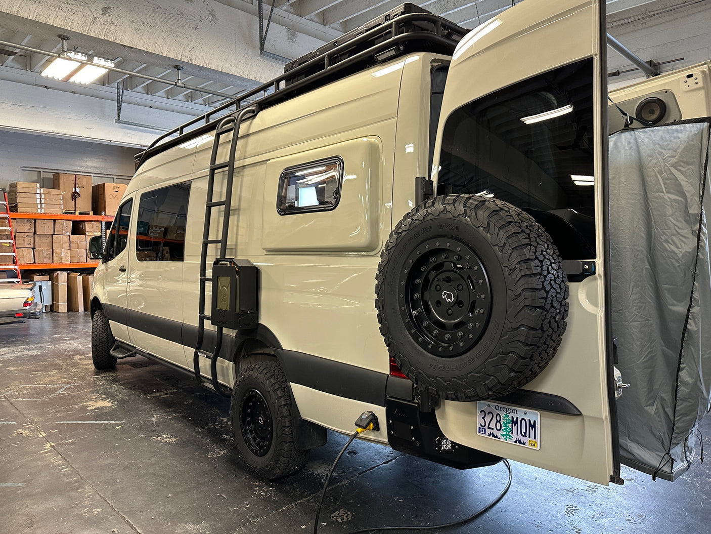 Customized RV rental van equipped for adventure: off-road tires, roof rack, side ladder, and rear outdoor shower.