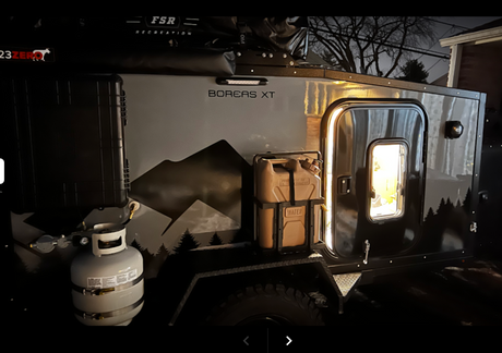 Night view of a gray Boreas XT overland camper with propane, water jug, & lighted door, great for RV rental.