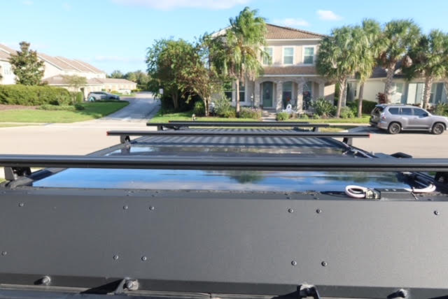 Roof-level view of the camper showing the mounted solar panel wired in, Alu-Cab 1450mm standard-profile load bars and the Sherpa Little Cub rack ahead of it — photo 13