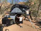 Rooftop tent camping with an RV rental! A teardrop trailer and ladder are ready for adventure. Two people smile in the desert.