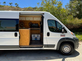 This white Ram ProMaster RV rental, with its side door open, reveals a cozy wooden interior kitchen, ready for adventure.