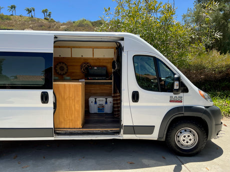 This white Ram ProMaster RV rental, with its side door open, reveals a cozy wooden interior kitchen, ready for adventure.
