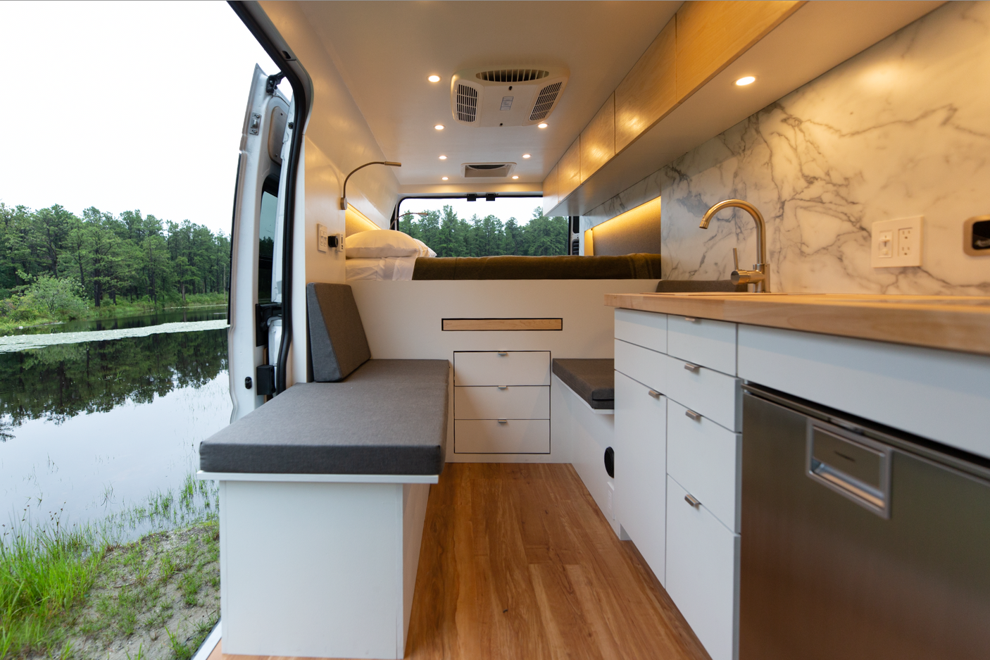 A spacious, modern RV rental camper van interior. The open rear doors reveal a serene lake with trees. Inside are a kitchen, elevated bed, and seating.