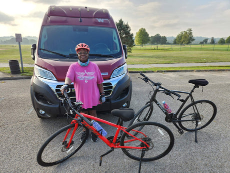A woman in a pink "Beat Cancer" shirt, bike helmet, and gloves stands by a red RV rental with two bicycles.