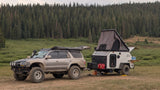 An off-road SUV tows an overlanding trailer with a rooftop tent, parked in a lush forest for a wilderness rv rental adventure.