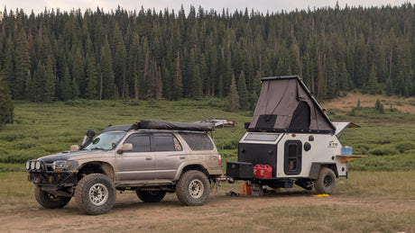 An off-road SUV tows an overlanding trailer with a rooftop tent, parked in a lush forest for a wilderness rv rental adventure.