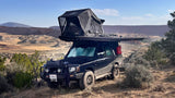 Black Land Rover SUV with rooftop tent & awning set up on a dirt path in a scenic desert landscape, like an rv rental.