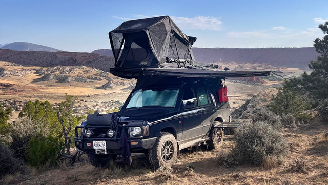 Black Land Rover SUV with rooftop tent & awning set up on a dirt path in a scenic desert landscape, like an rv rental.