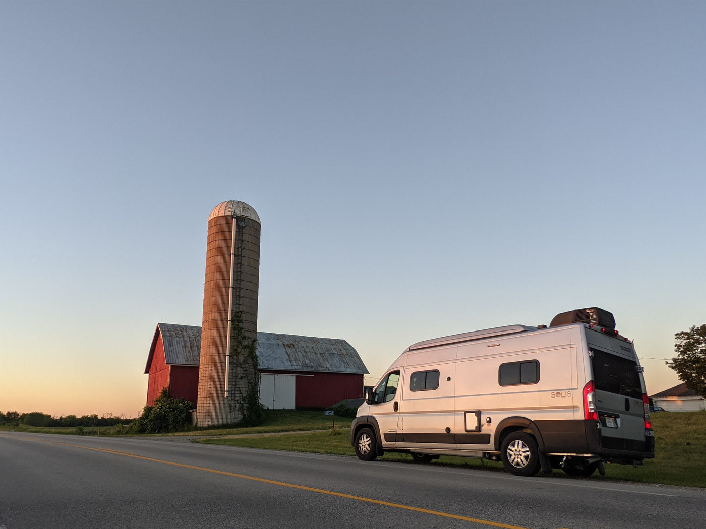 An RV rental camper van parked roadside by a farm, featuring a red barn and silo, under a golden hour sky. — photo 17
