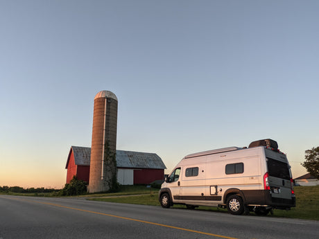 An RV rental camper van parked roadside by a farm, featuring a red barn and silo, under a golden hour sky.