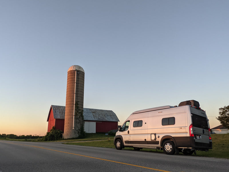 An RV rental camper van parked roadside by a farm, featuring a red barn and silo, under a golden hour sky. — photo 17