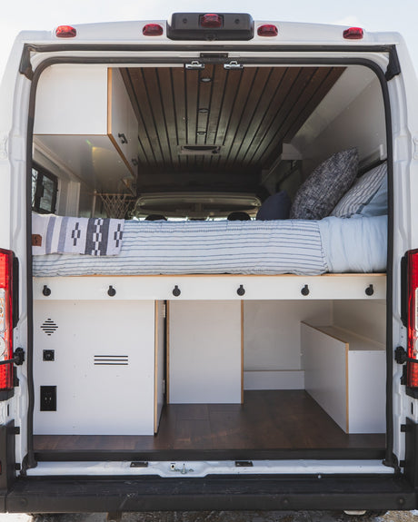 Rear view of a white camper van interior with a bed, under-bed storage, and wood ceiling. An ideal rv rental setup for travel.