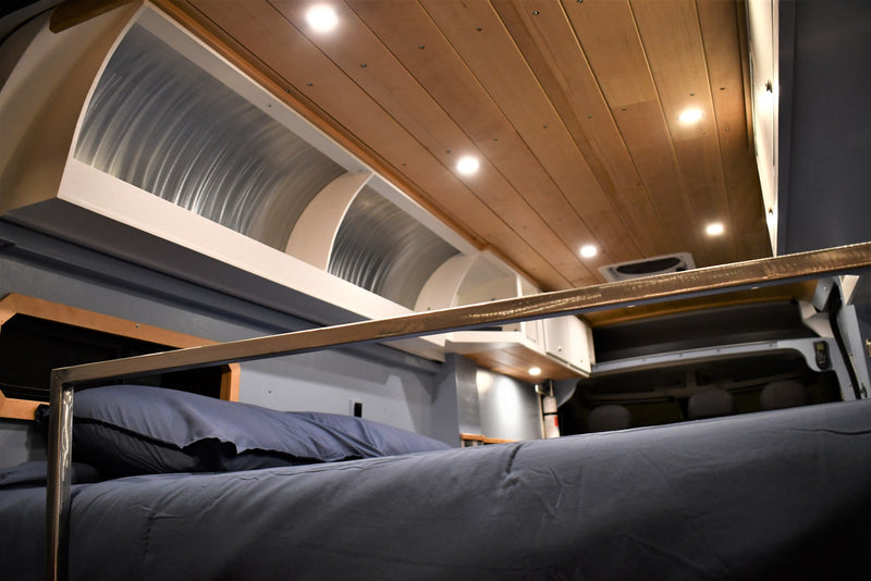 Inside an RV rental: a cozy bed with dark bedding, metal bunk rail, unique curved overhead storage, and wood ceiling. — photo 13