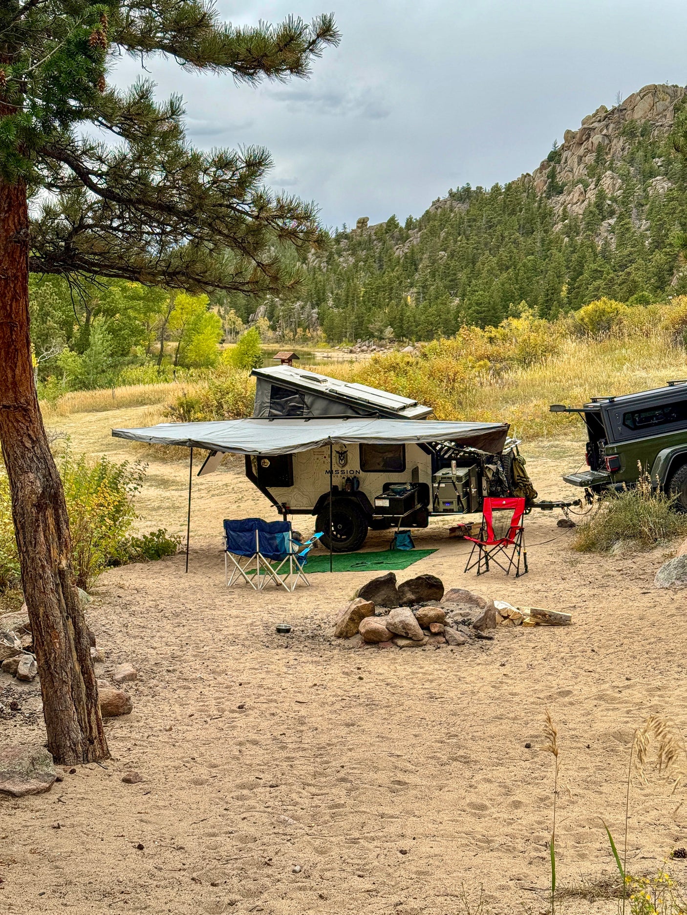 Enjoy nature with this overland trailer, an RV rental option, parked in a sandy campsite with chairs, fire pit, and mountain views.