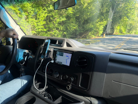 RV rental drive: Navigation on dashboard screen and phone mount. Driver's view of green trees and road ahead.