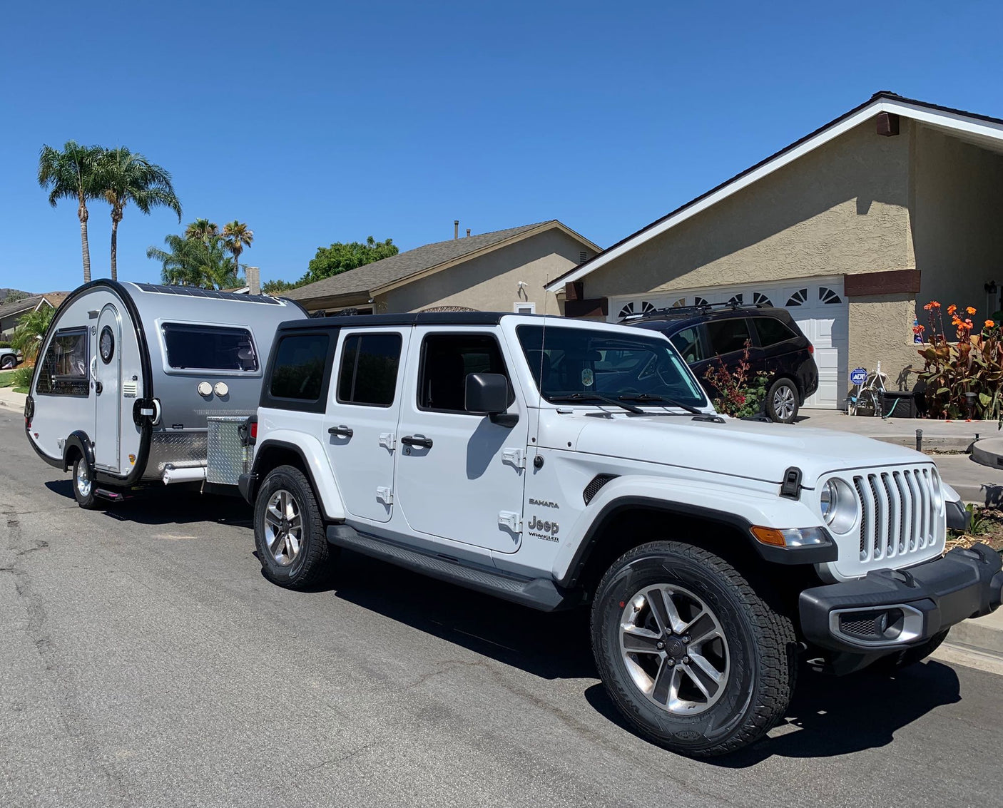 This white Jeep Wrangler tows a modern teardrop RV rental, parked on a residential street under a blue sky.
