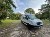 White Mercedes-Benz Sprinter van RV rental parked on a dirt path with trees. Kayaks and grill visible in background.