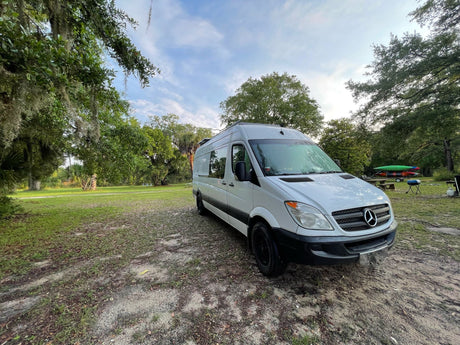 White Mercedes-Benz Sprinter van RV rental parked on a dirt path with trees. Kayaks and grill visible in background.