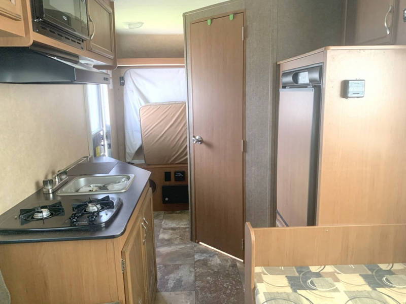 Inside an RV rental: a compact kitchen (stove, sink, microwave), fridge, and bed area, designed for efficient living.
