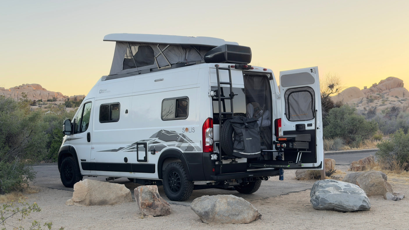 This white RV rental camper van, with its pop-top roof and outdoor kitchen, is parked in a scenic desert landscape at dusk, ready for adventure.