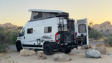 This white RV rental camper van, with its pop-top roof and outdoor kitchen, is parked in a scenic desert landscape at dusk, ready for adventure.