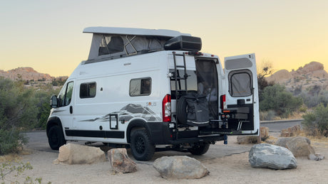 This white RV rental camper van, with its pop-top roof and outdoor kitchen, is parked in a scenic desert landscape at dusk, ready for adventure.