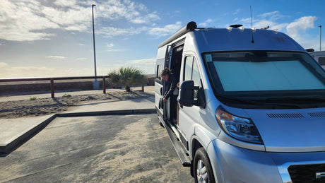 A person leans out of the side door of a silver RV rental parked by the beach, enjoying the ocean view on a sunny day.