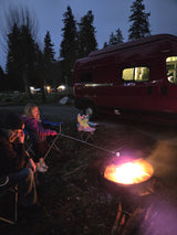 Family camping at dusk: A child roasts marshmallows by the campfire with an adult, next to their red RV rental camper van.